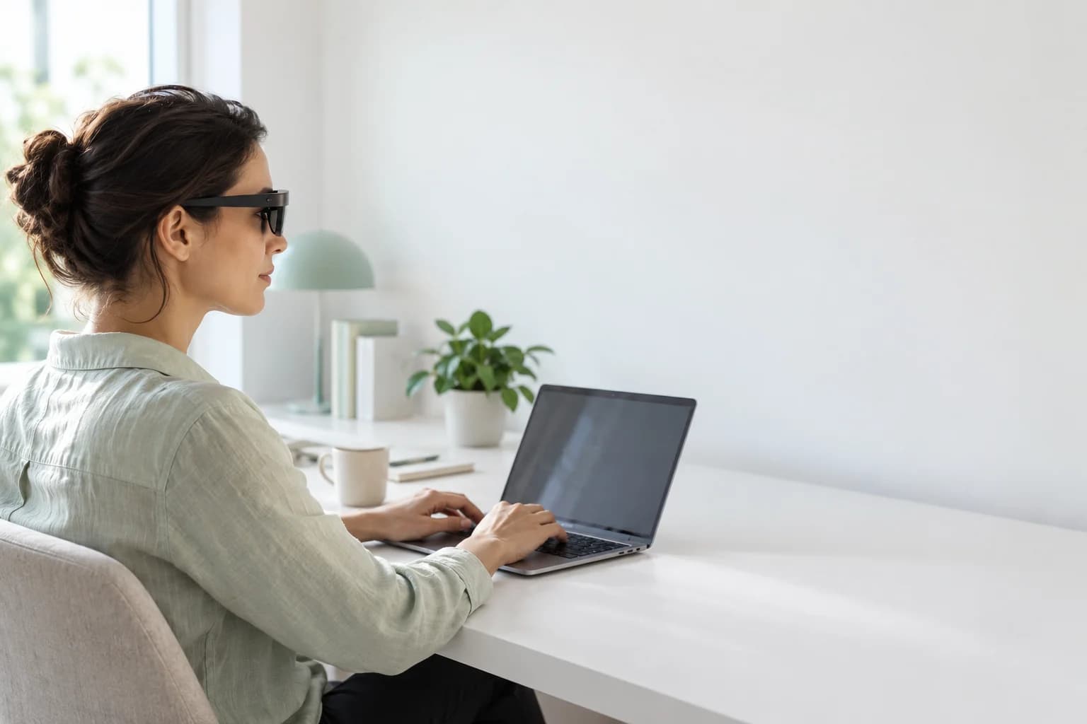 Eine Frau trägt eine AR-Brille am Schreibtisch und blickt über ihren Laptop.